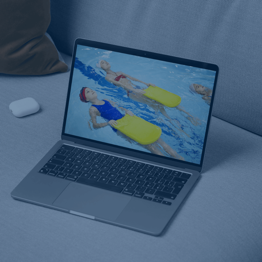 Ein Laptop auf einer Couch zeigt Kinder, die mit gelben Schwimmbrettern im Wasser trainieren – ein Einblick in die AquaFit Schwimmakademie und ihre Online-Kursangebote.