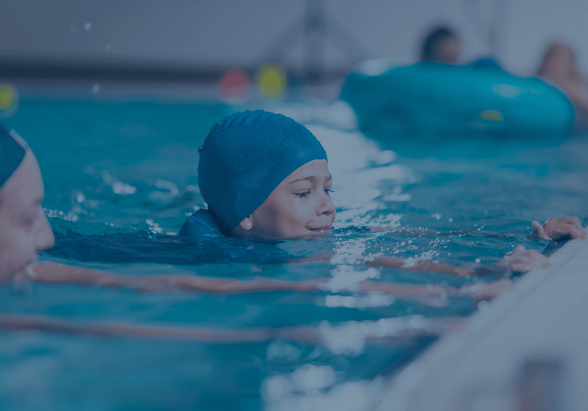 Ein Kind mit blauer Badekappe übt im Schwimmbecken Beinbewegungen und hält sich am Beckenrand fest, während ein Trainer unterstützend daneben ist.