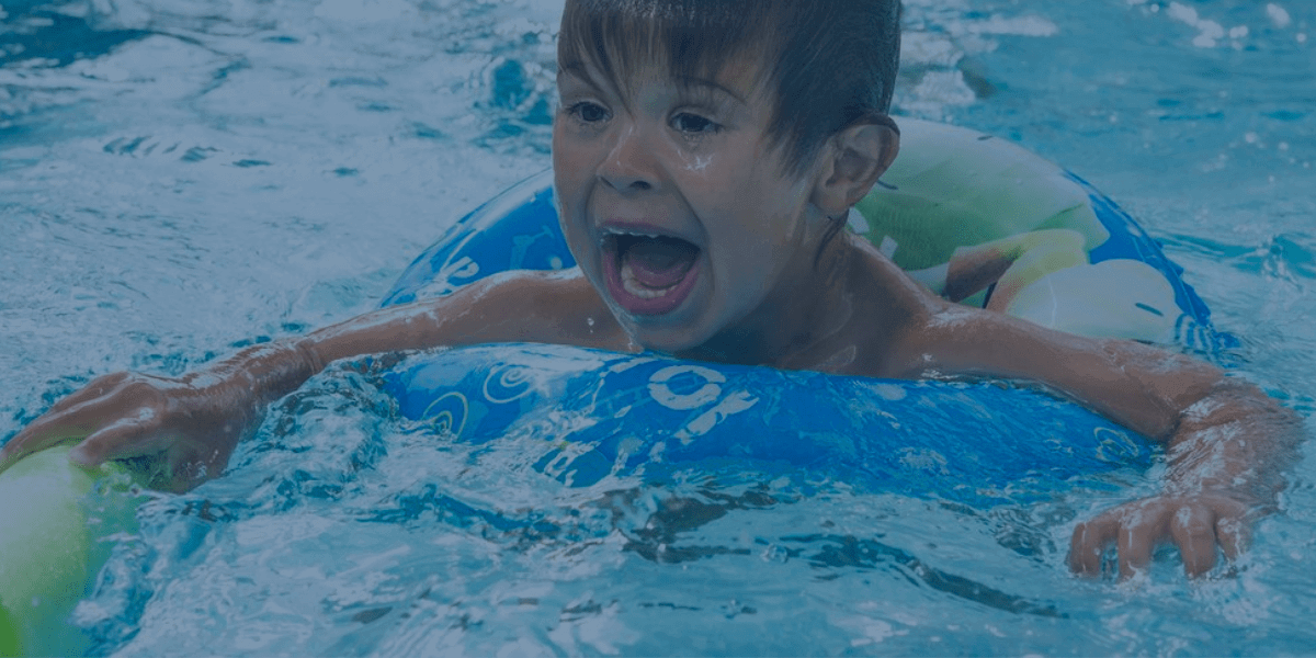 Ein Kind in einem Schwimmkurs hält sich an einem Schwimmbrett fest, während es mit Unterstützung seine Wasserängste überwindet.