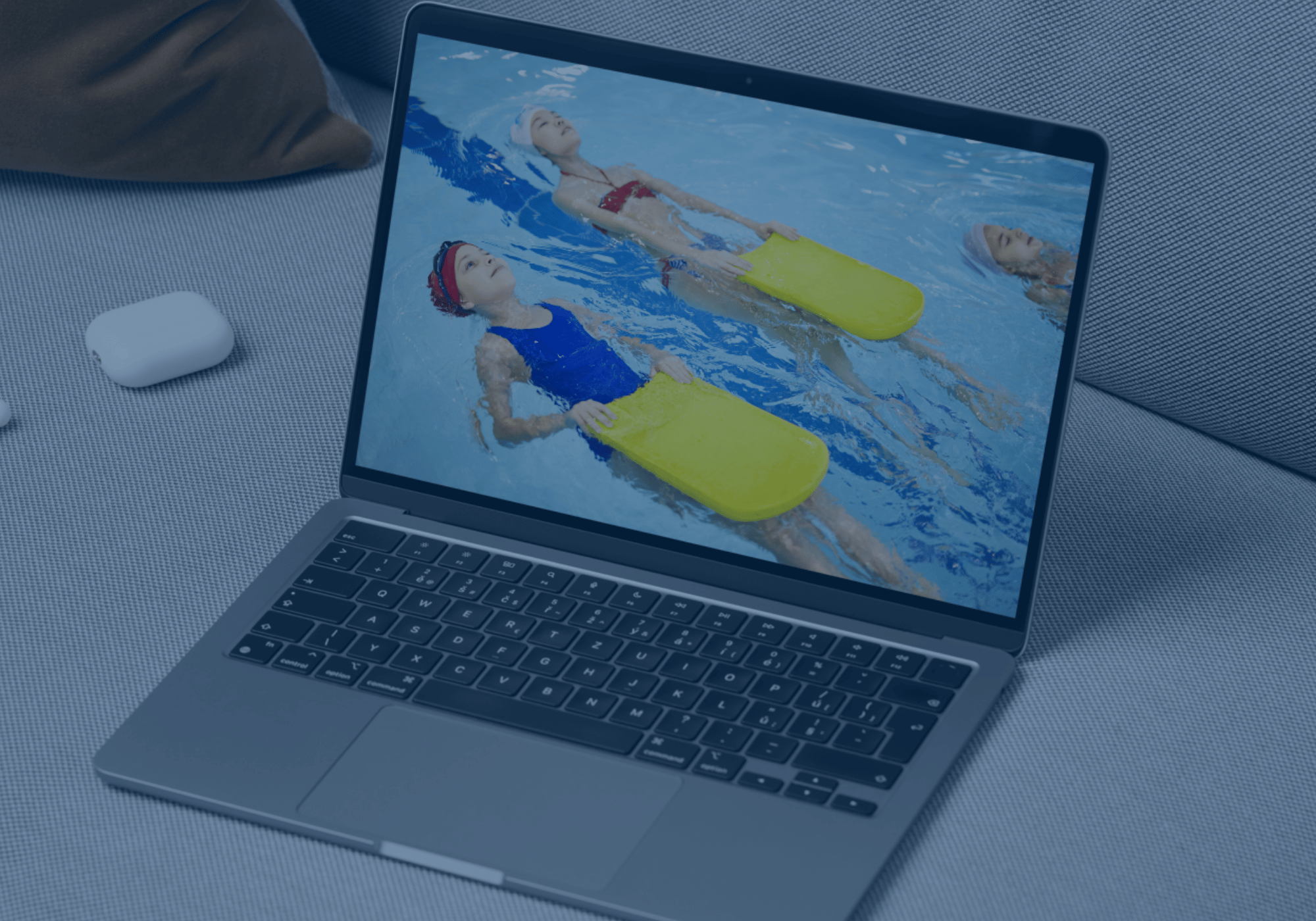 Ein Laptop auf einer Couch zeigt Kinder, die mit gelben Schwimmbrettern im Wasser trainieren – ein Einblick in die AquaFit Schwimmakademie und ihre Online-Kursangebote.