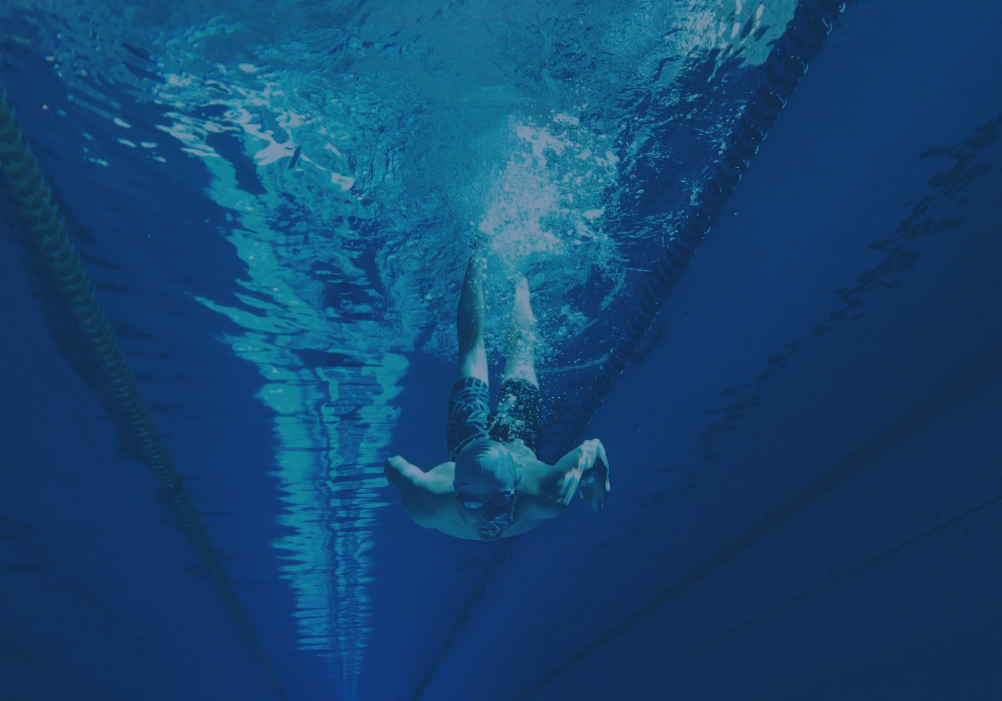 Unterwasseraufnahme eines Schwimmers in einem blauen Pool, der sich kraftvoll vom Beckenrand abstößt – Symbol für Geschwindigkeit und Technik.